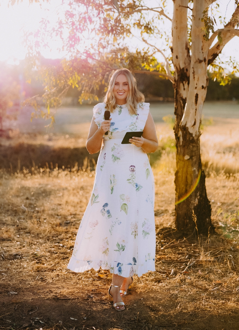 Cassie Argus, a professional and warmly presented celebrant and MC, holding a microphone ready to host a joyful milestone celebration.