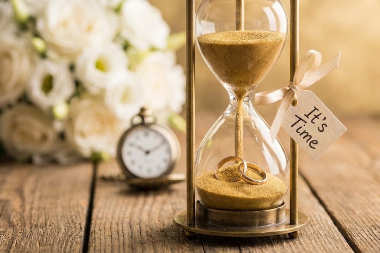 A traditional hourglass with an 'it's time' tag alongside a fob watch and flowers, representing the countdown to a beautiful Heathcote wedding.